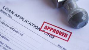 Close-up of a loan application form stamped with "APPROVED" in red ink, symbolizing successful business loan qualification. A gray rubber stamp is placed on the document, emphasizing loan approval.